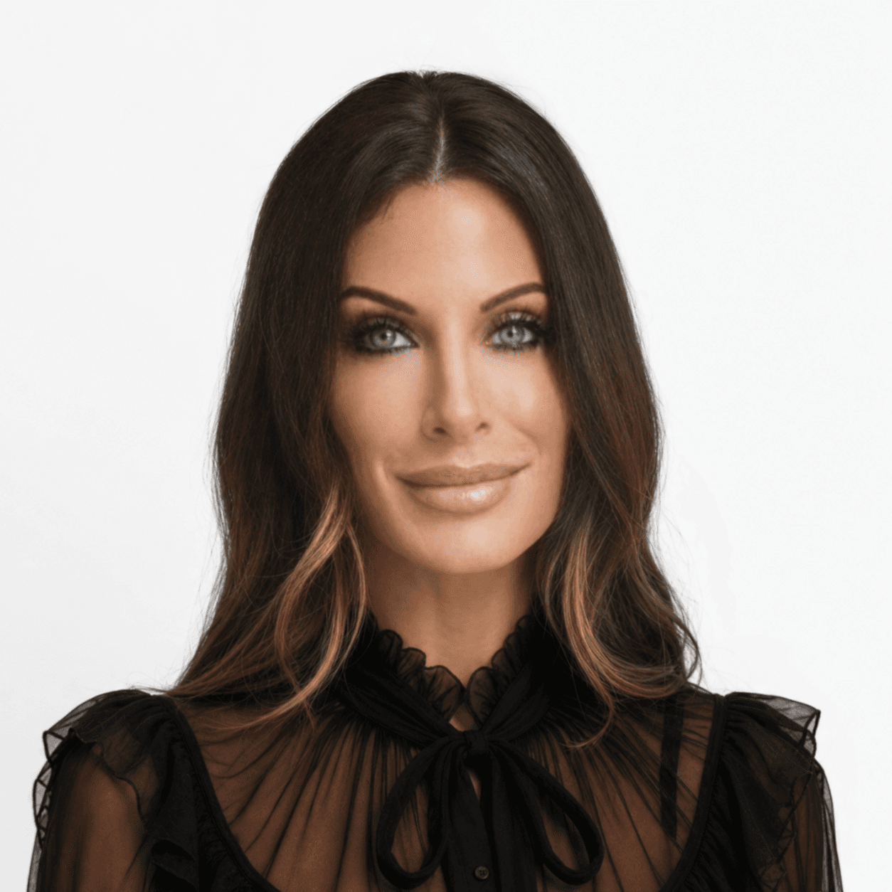 Smiling woman with long dark hair, light eyes, dark eye makeup, and a black sheer blouse with a front bow.